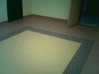 Wykończona podłoga w pomieszczeniu: jasna, kwadratowa powierzchnia z obramowaniem z ciemniejszego, nakrapianego materiału. Widoczne listwy przypodłogowe i fragment otwartych drzwi.